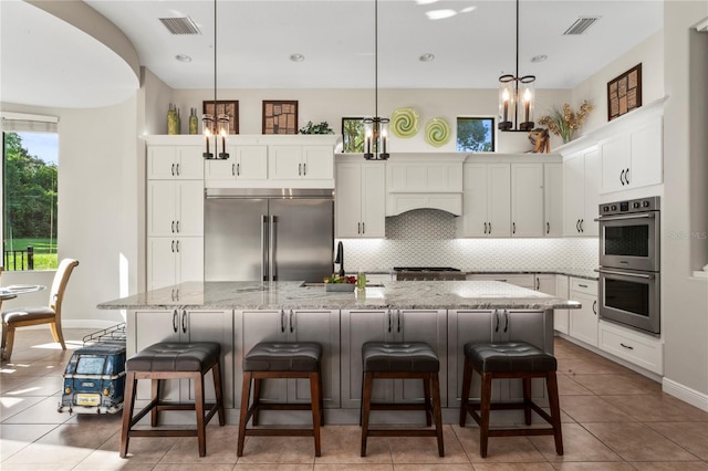 kitchen featuring white cabinetry, tasteful backsplash, a kitchen island with sink, stainless steel appliances, and decorative light fixtures