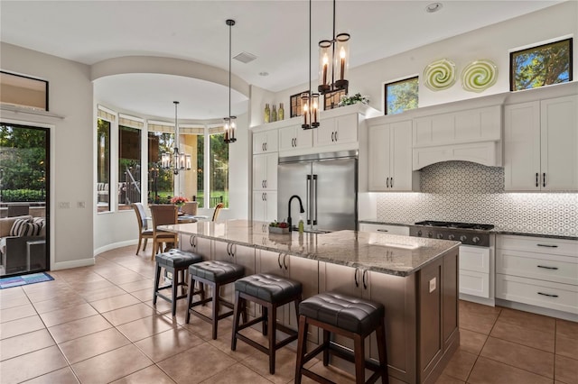 kitchen with white cabinetry, stainless steel built in refrigerator, a kitchen island with sink, decorative backsplash, and pendant lighting