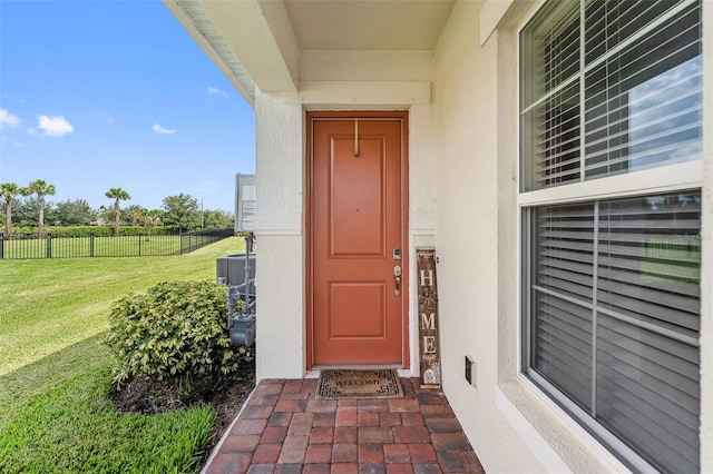 entrance to property featuring fence, a lawn, central AC unit, and stucco siding