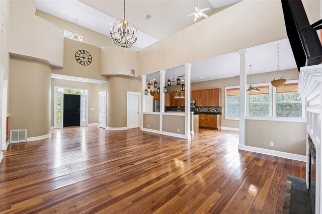 unfurnished living room with ceiling fan with notable chandelier, a towering ceiling, ornate columns, and dark hardwood / wood-style flooring