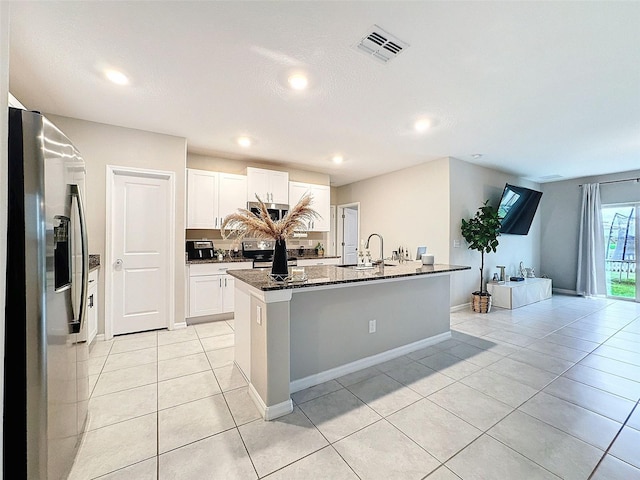 kitchen featuring a center island with sink, light tile patterned floors, stainless steel appliances, white cabinetry, and dark stone countertops