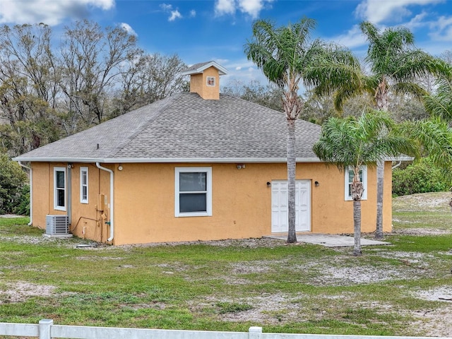 rear view of house with a yard, central AC unit, stucco siding, and roof with shingles