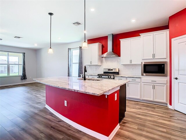 kitchen with hardwood / wood-style floors, appliances with stainless steel finishes, white cabinetry, wall chimney range hood, and a kitchen island with sink