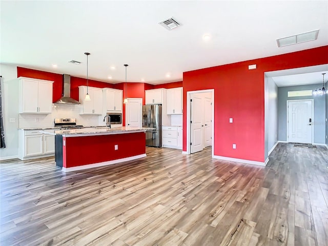 kitchen featuring white cabinets, appliances with stainless steel finishes, wood-type flooring, wall chimney exhaust hood, and a kitchen island with sink