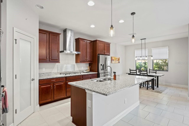 kitchen featuring an island with sink, a sink, decorative light fixtures, stainless steel appliances, and wall chimney exhaust hood