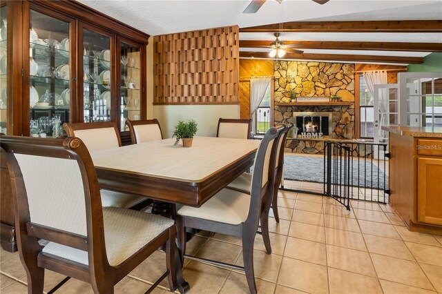 dining area with a fireplace, wood walls, ceiling fan, beam ceiling, and light tile patterned flooring