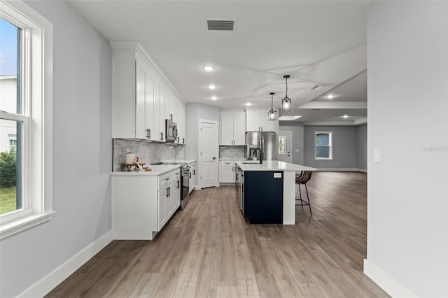 kitchen with pendant lighting, light hardwood / wood-style floors, an island with sink, white cabinetry, and appliances with stainless steel finishes