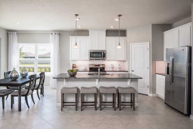 kitchen with appliances with stainless steel finishes, dark stone countertops, white cabinetry, an island with sink, and pendant lighting