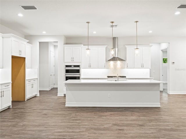 kitchen featuring decorative light fixtures, light hardwood / wood-style flooring, a center island with sink, and wall chimney range hood