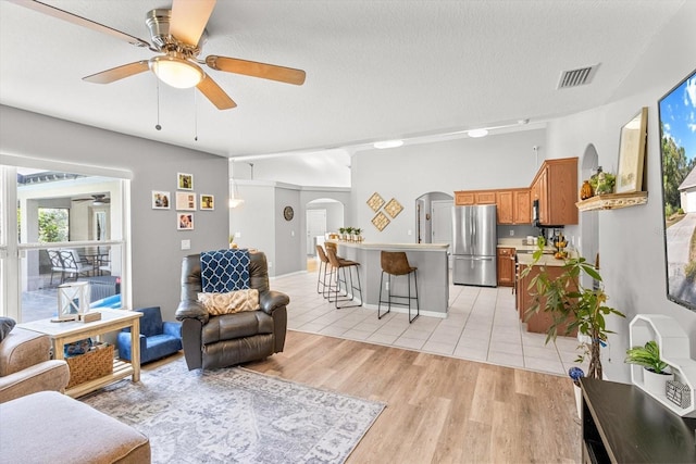 living room with arched walkways, visible vents, light wood-style floors, ceiling fan, and a textured ceiling