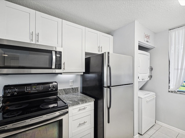 kitchen featuring stacked washing maching and dryer, dark stone counters, stainless steel appliances, light tile patterned floors, and white cabinetry