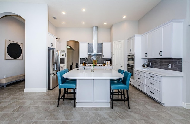 kitchen with a kitchen bar, wall chimney range hood, an island with sink, and appliances with stainless steel finishes