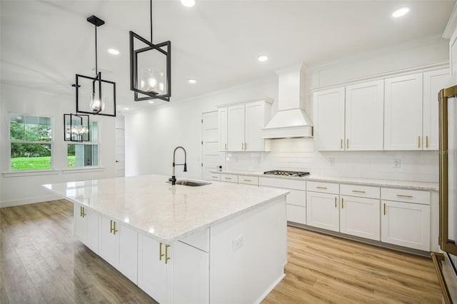 kitchen with white cabinets, an island with sink, premium range hood, light wood-type flooring, and sink