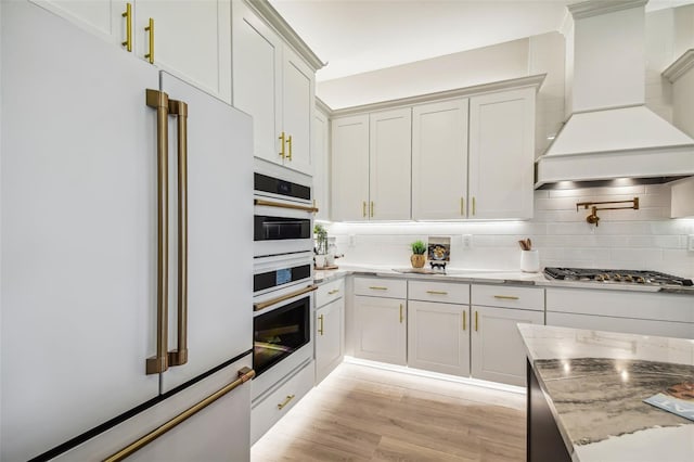 kitchen with light wood-style flooring, backsplash, light stone countertops, white appliances, and premium range hood