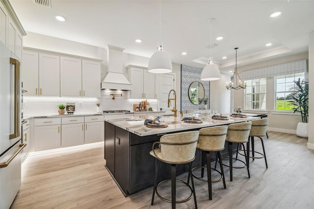 kitchen featuring light stone counters, a center island with sink, custom exhaust hood, a raised ceiling, and hanging light fixtures