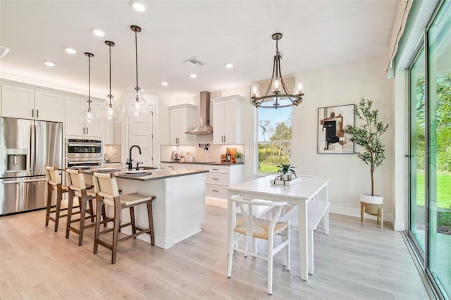 kitchen with an island with sink, white cabinets, pendant lighting, wall chimney range hood, and appliances with stainless steel finishes