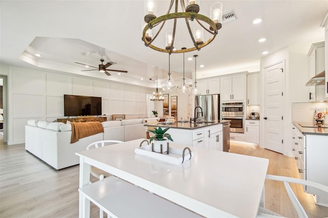 kitchen with a raised ceiling, hanging light fixtures, a kitchen island with sink, and light wood-type flooring