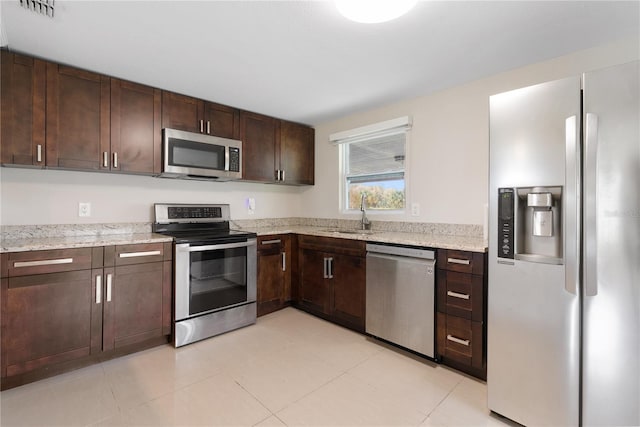 kitchen with appliances with stainless steel finishes, sink, dark brown cabinets, light stone counters, and light tile patterned floors