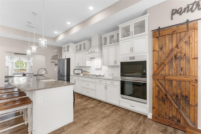 kitchen with a center island with sink, custom exhaust hood, light stone countertops, dark hardwood / wood-style floors, and appliances with stainless steel finishes