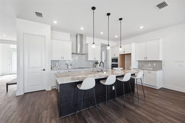 kitchen with wall chimney range hood, white cabinetry, stainless steel appliances, dark hardwood / wood-style floors, and a center island with sink