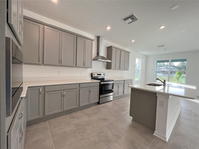 kitchen with wall chimney exhaust hood, an island with sink, sink, gray cabinets, and appliances with stainless steel finishes