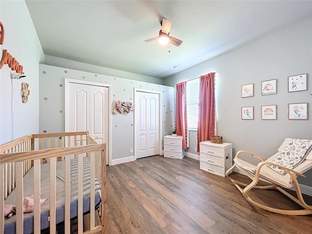 bedroom with dark wood-type flooring, ceiling fan, two closets, and a nursery area