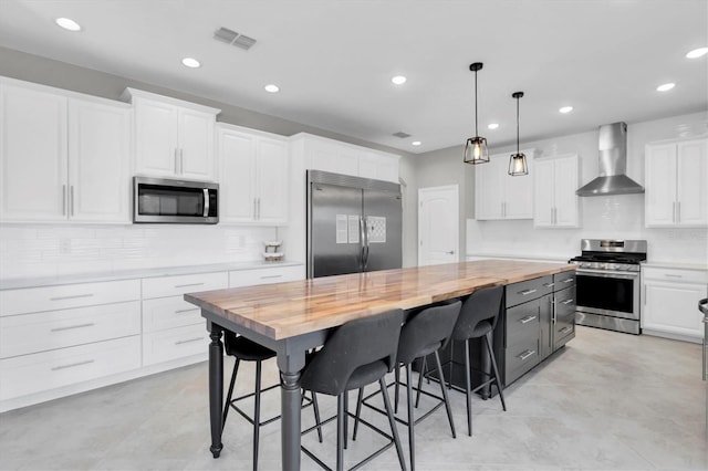 kitchen featuring wall chimney range hood, white cabinets, a kitchen island, appliances with stainless steel finishes, and wooden counters