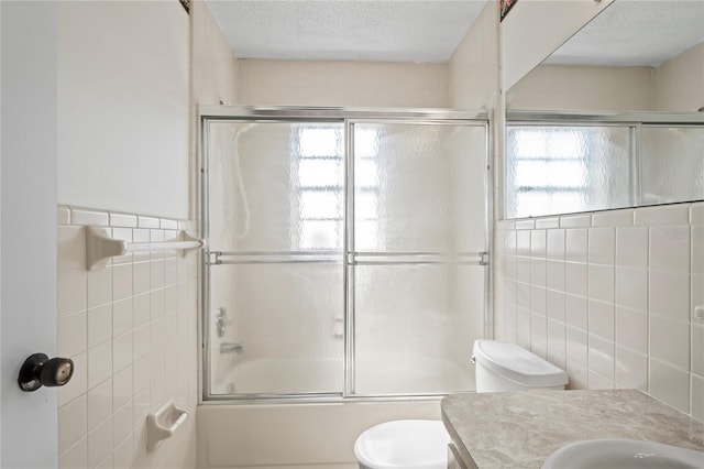 full bathroom featuring vanity, shower / bath combination with glass door, toilet, a textured ceiling, and tile walls