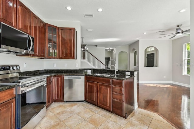 kitchen with stainless steel appliances, dark stone counters, sink, light wood-type flooring, and ceiling fan with notable chandelier