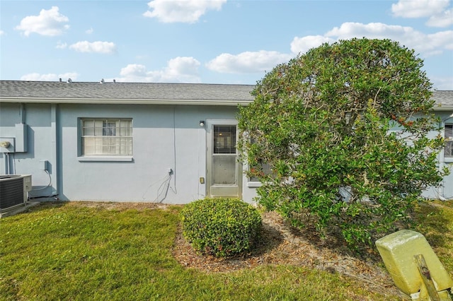 rear view of house with central air condition unit, stucco siding, a yard, and a shingled roof
