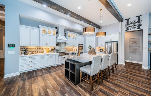 kitchen with beamed ceiling, custom exhaust hood, white cabinetry, and an island with sink