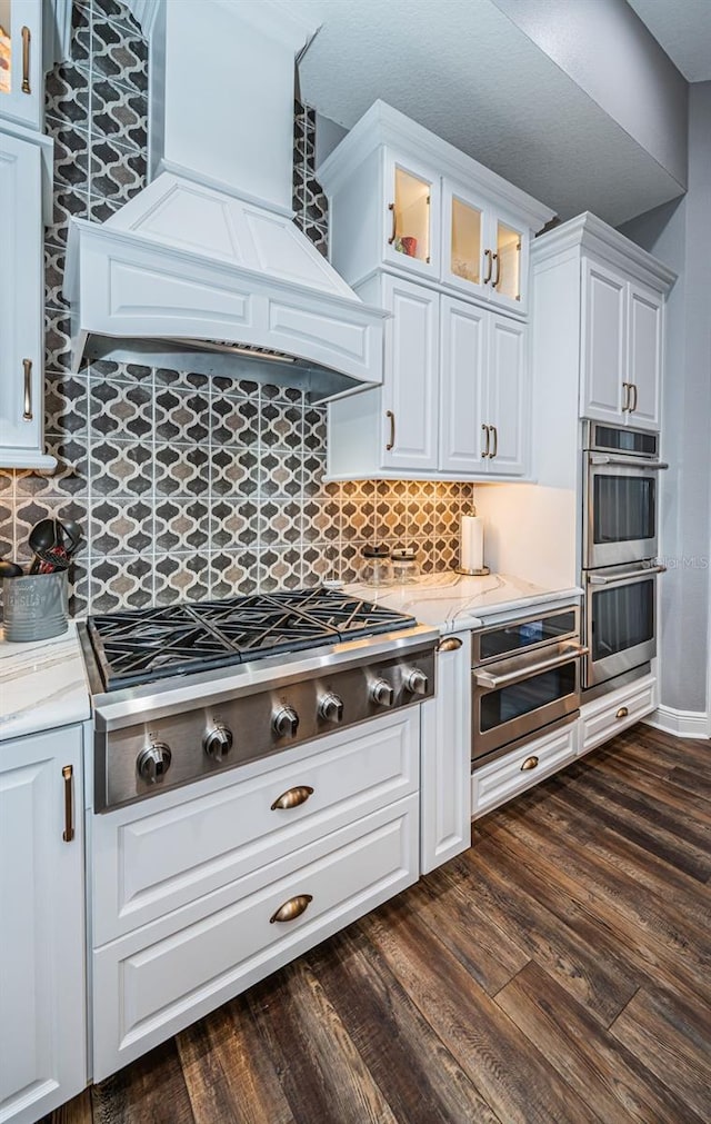 kitchen featuring dark wood-type flooring, custom range hood, stainless steel appliances, backsplash, and white cabinetry