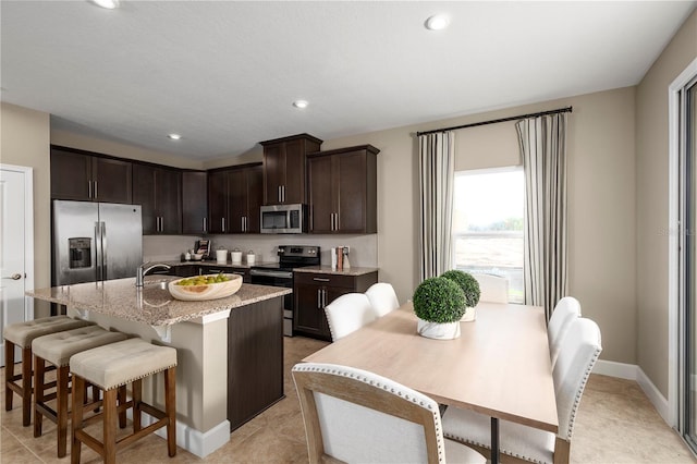 kitchen with light stone countertops, a kitchen bar, an island with sink, stainless steel appliances, and dark brown cabinetry