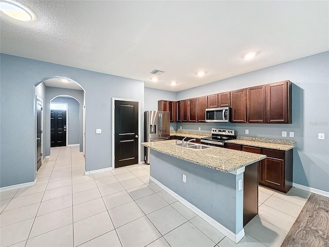 kitchen with stainless steel appliances, light stone counters, a textured ceiling, a kitchen island with sink, and light tile patterned floors