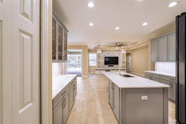 kitchen featuring sink, a tray ceiling, gray cabinets, an island with sink, and ceiling fan