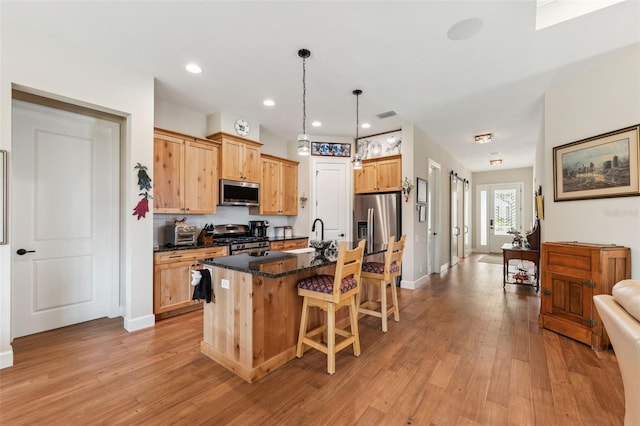 kitchen featuring light hardwood / wood-style floors, a center island with sink, dark stone counters, appliances with stainless steel finishes, and decorative light fixtures