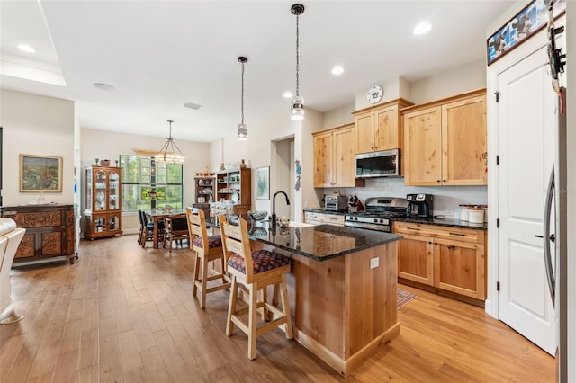 kitchen with stainless steel appliances, an island with sink, pendant lighting, light hardwood / wood-style flooring, and dark stone countertops