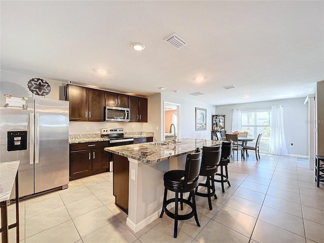 kitchen with appliances with stainless steel finishes, light stone counters, sink, light tile patterned floors, and a center island with sink