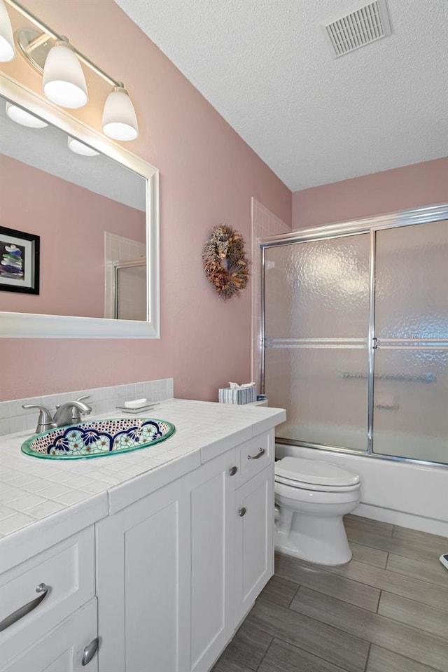 full bathroom with vanity, bath / shower combo with glass door, a textured ceiling, and toilet