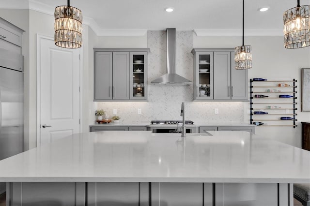 kitchen with gray cabinetry, a large island with sink, wall chimney range hood, hanging light fixtures, and ornamental molding