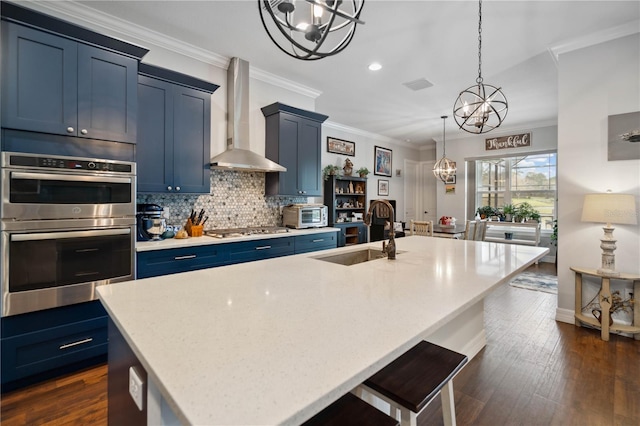 kitchen featuring appliances with stainless steel finishes, blue cabinets, sink, wall chimney range hood, and an island with sink