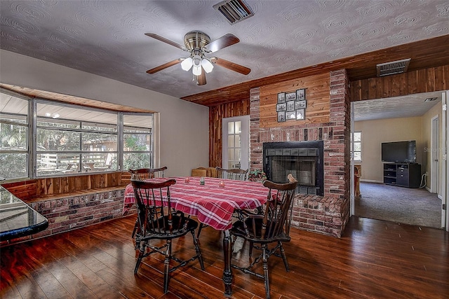 dining space featuring ceiling fan, a brick fireplace, dark hardwood / wood-style flooring, a textured ceiling, and wooden walls