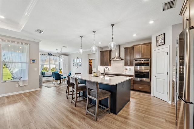 kitchen with appliances with stainless steel finishes, light wood-type flooring, an island with sink, and wall chimney range hood