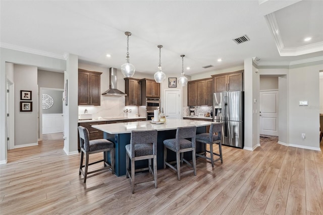 kitchen featuring a spacious island, wall chimney range hood, hanging light fixtures, light hardwood / wood-style flooring, and stainless steel appliances