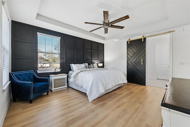 bedroom featuring a raised ceiling, a barn door, ceiling fan, and light wood-type flooring