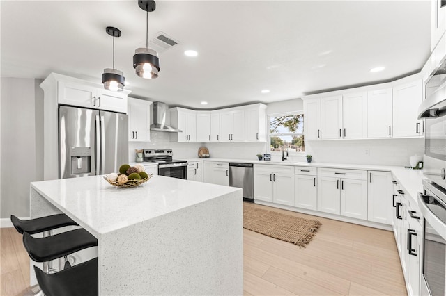 kitchen featuring pendant lighting, wall chimney range hood, light wood-type flooring, appliances with stainless steel finishes, and white cabinetry