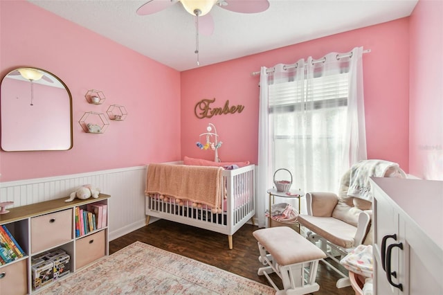 bedroom with a crib, ceiling fan, and dark wood-type flooring