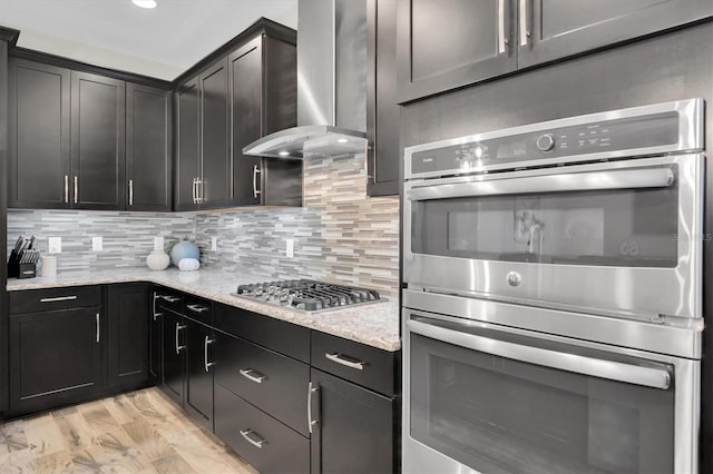 kitchen featuring stainless steel appliances, decorative backsplash, light stone countertops, wall chimney exhaust hood, and light hardwood / wood-style flooring