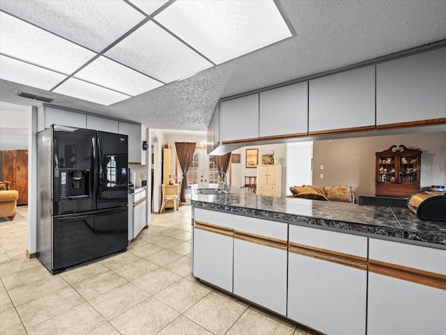 kitchen with kitchen peninsula, light tile patterned floors, a textured ceiling, black fridge with ice dispenser, and white cabinetry