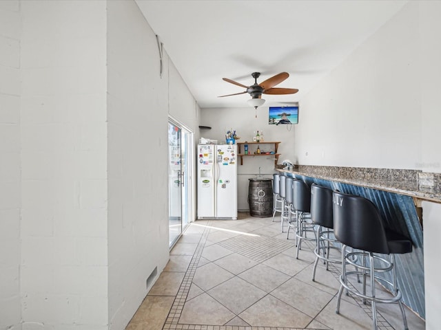 kitchen with white refrigerator with ice dispenser, light tile patterned floors, ceiling fan, and a kitchen breakfast bar